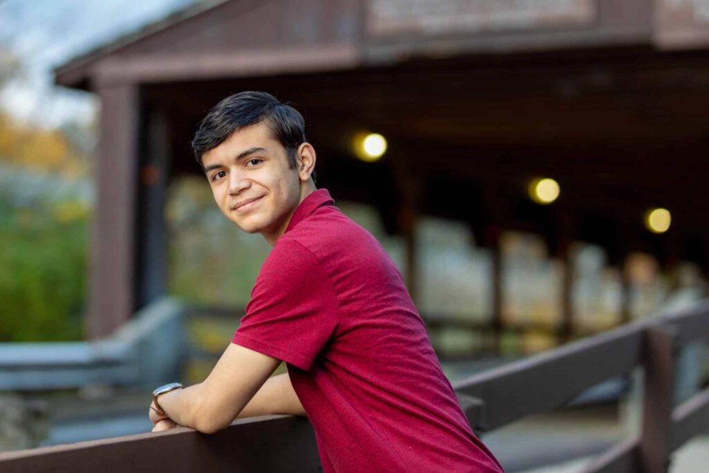 Alex in his senior picture, he is sitting in front of a covered bridge looking at camera