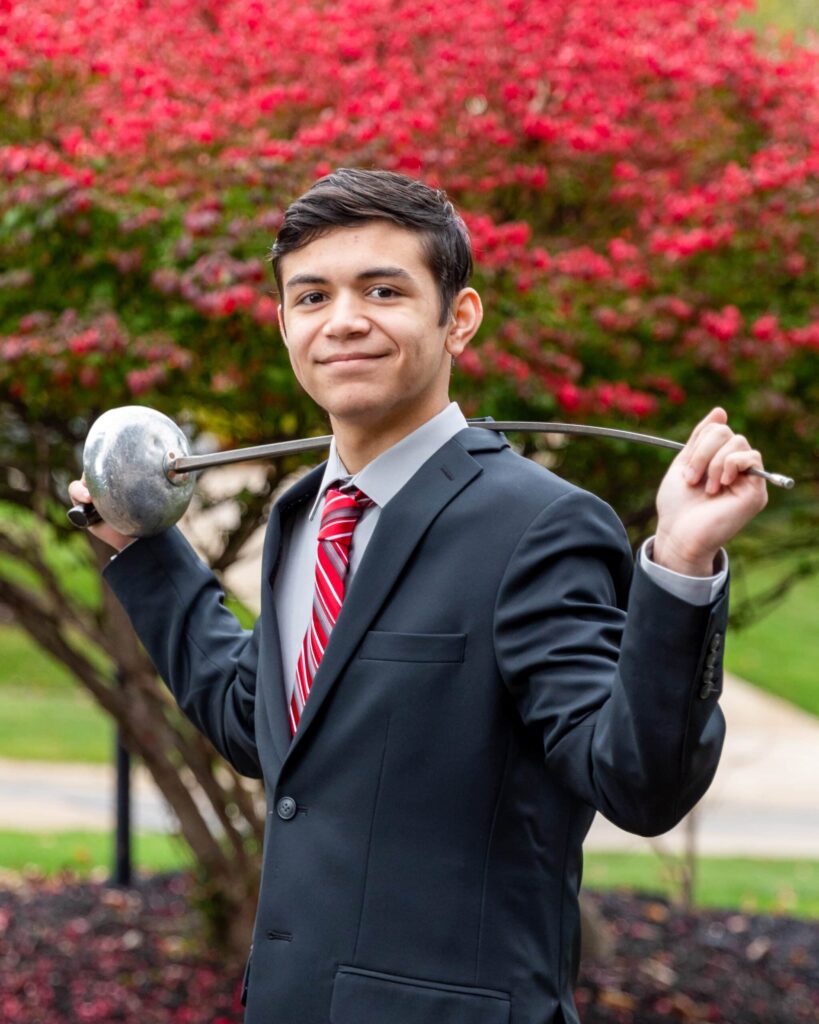 Alex in a suite and holding an epee behind his back