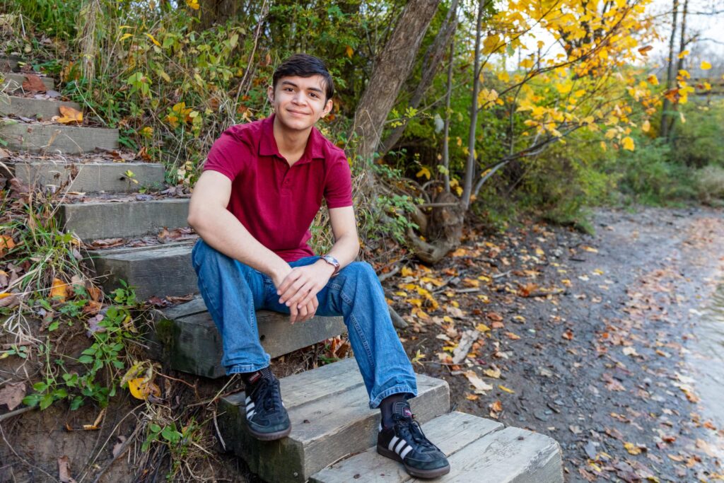 Alex in his senior portrait, wearing jeans and red polo shirt sitting on wooden stairs near a peaceful body of water